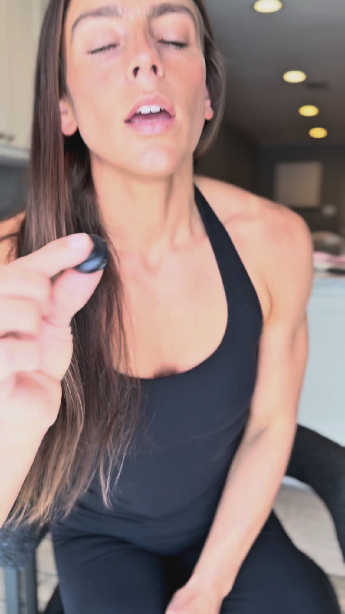 A woman wearing a black athletic tank top and matching leggings sits in a chair in a modern kitchen setting giving a testimonial about Noble Protein Powder